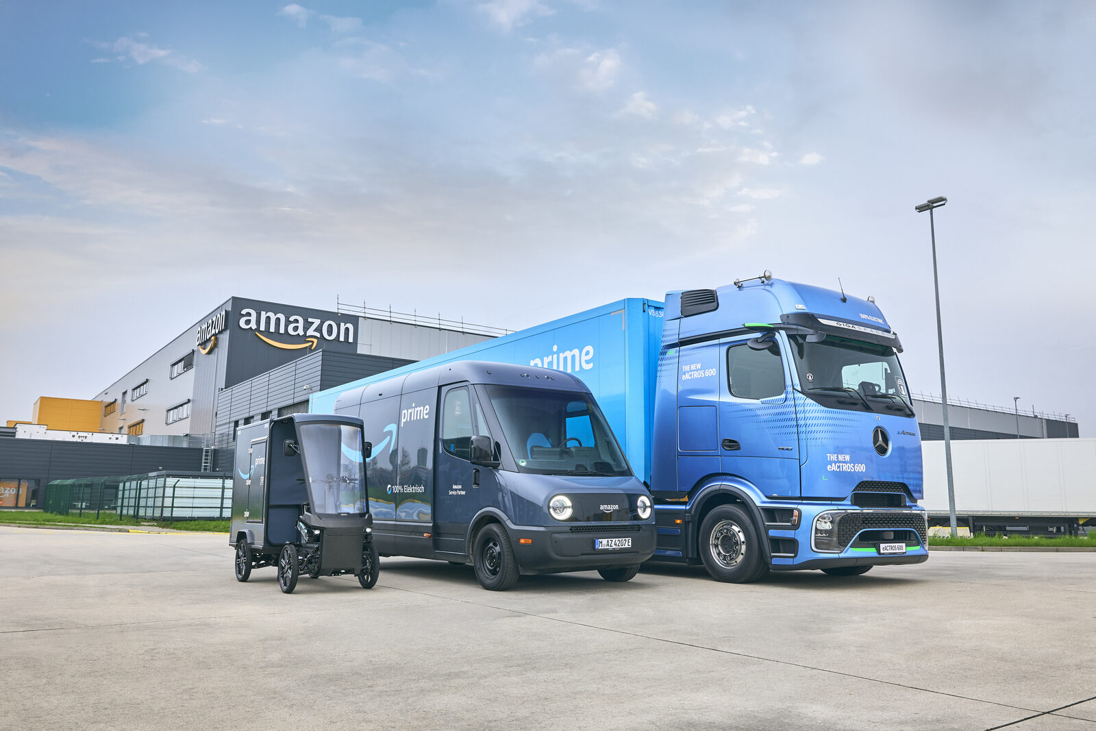Elektrische Lieferfahrzeuge und ein Lkw mit Prime-Logo vor einem Amazon-Logistikgebäude