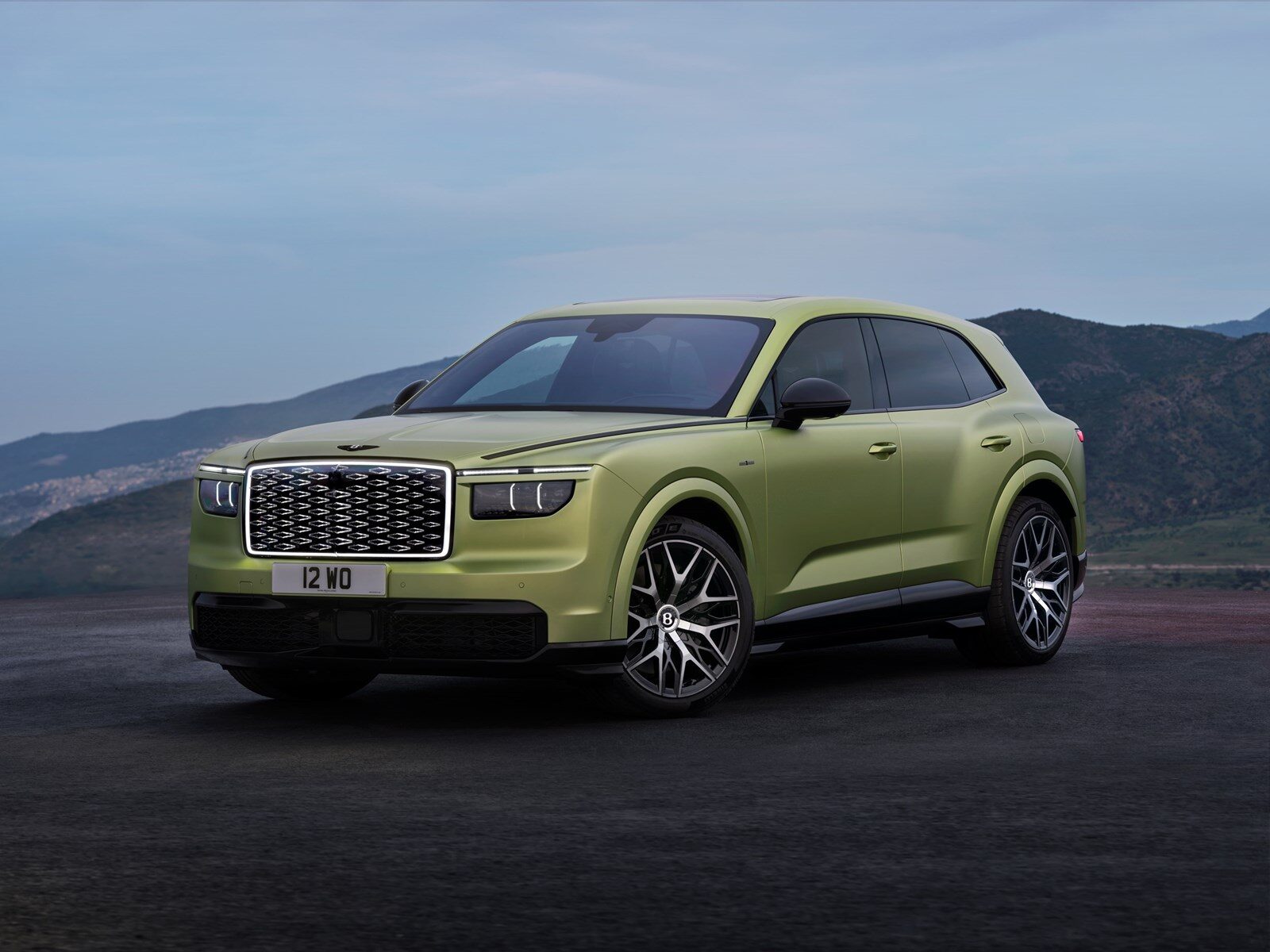 Yeşil renkli Bentley Torcal elektrikli SUV'nin ışıklı ızgaralı önden çapraz görünümü, arkada dağlar