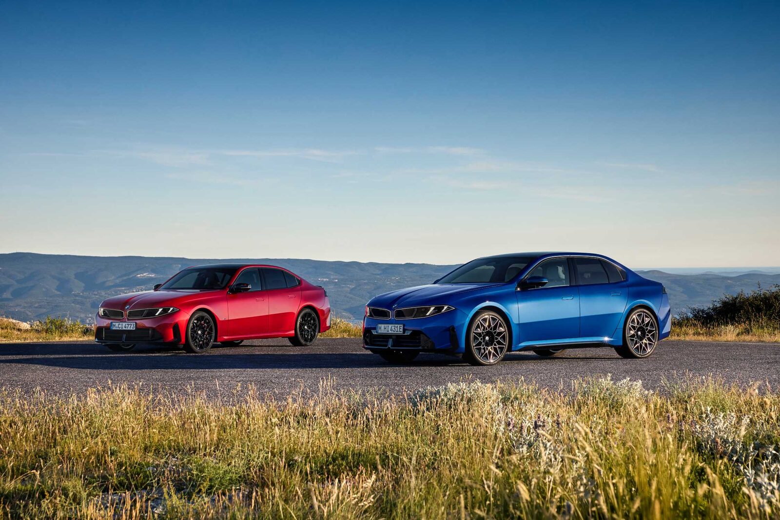 Yeni nesil BMW 3 Serisi'nin kırmızı ve mavi iki otomobili, tepelere bakan bir yol kenarında yan yana duruyor