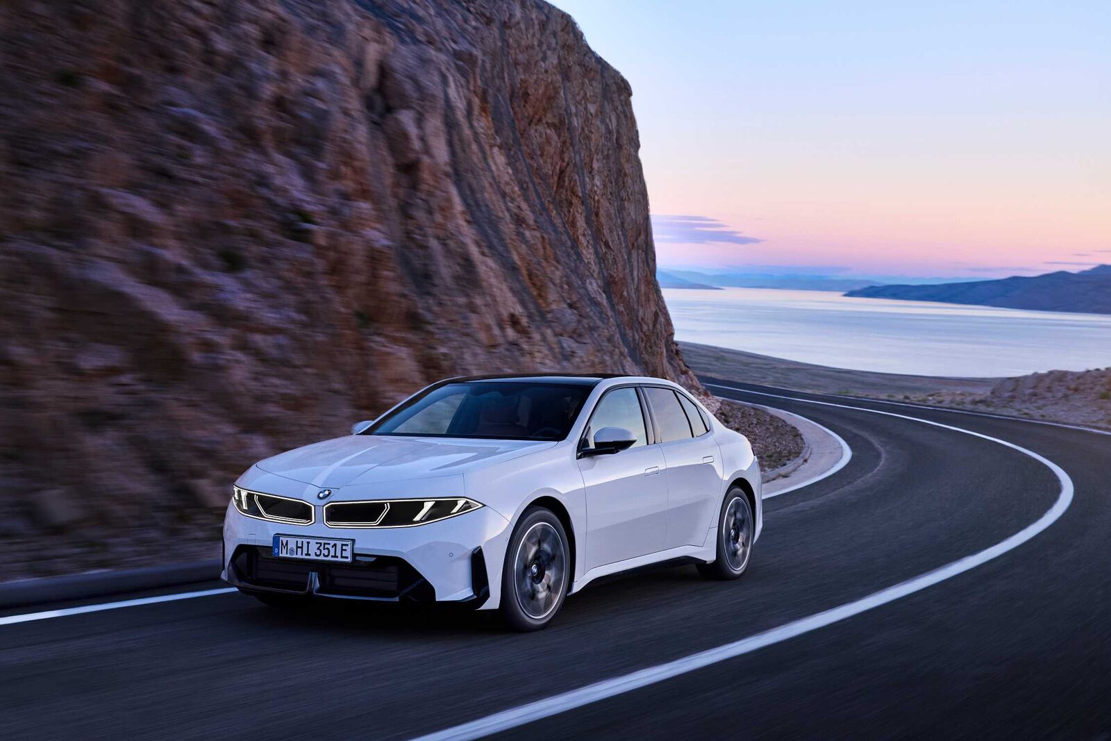Weißer BMW i3 der Neuen Klasse in einer Kurve an einer Felswand, im Hintergrund das Meer in der Abenddämmerung