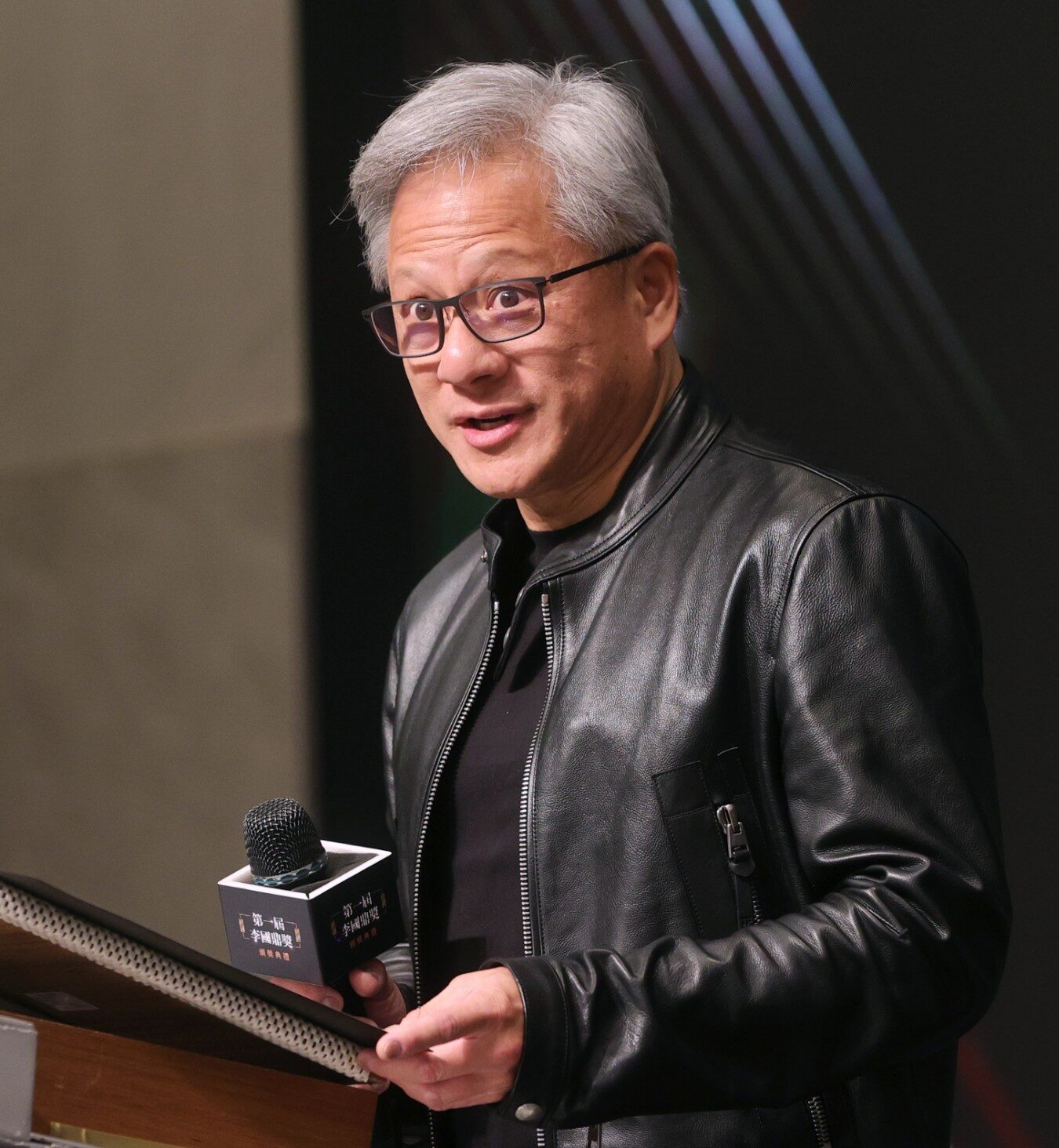 Siyah deri ceketli Jensen Huang elinde mikrofonla kürsüde konuşurken