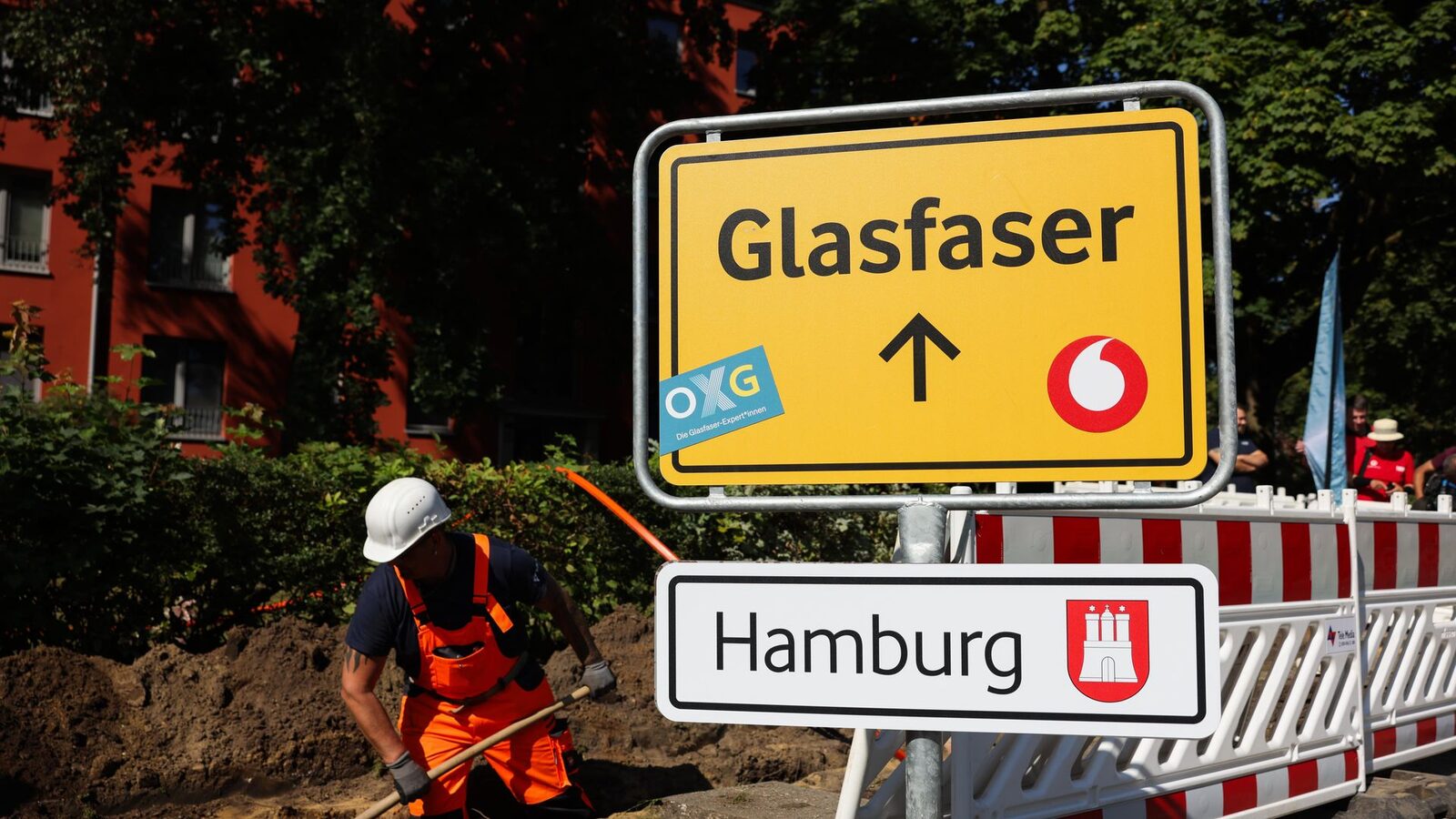 Bauarbeiter verlegt Glasfaser in einem Graben, daneben ein gelbes Schild „Glasfaser“ mit OXG- und Vodafone-Logo über einem Ortsschild von Hamburg