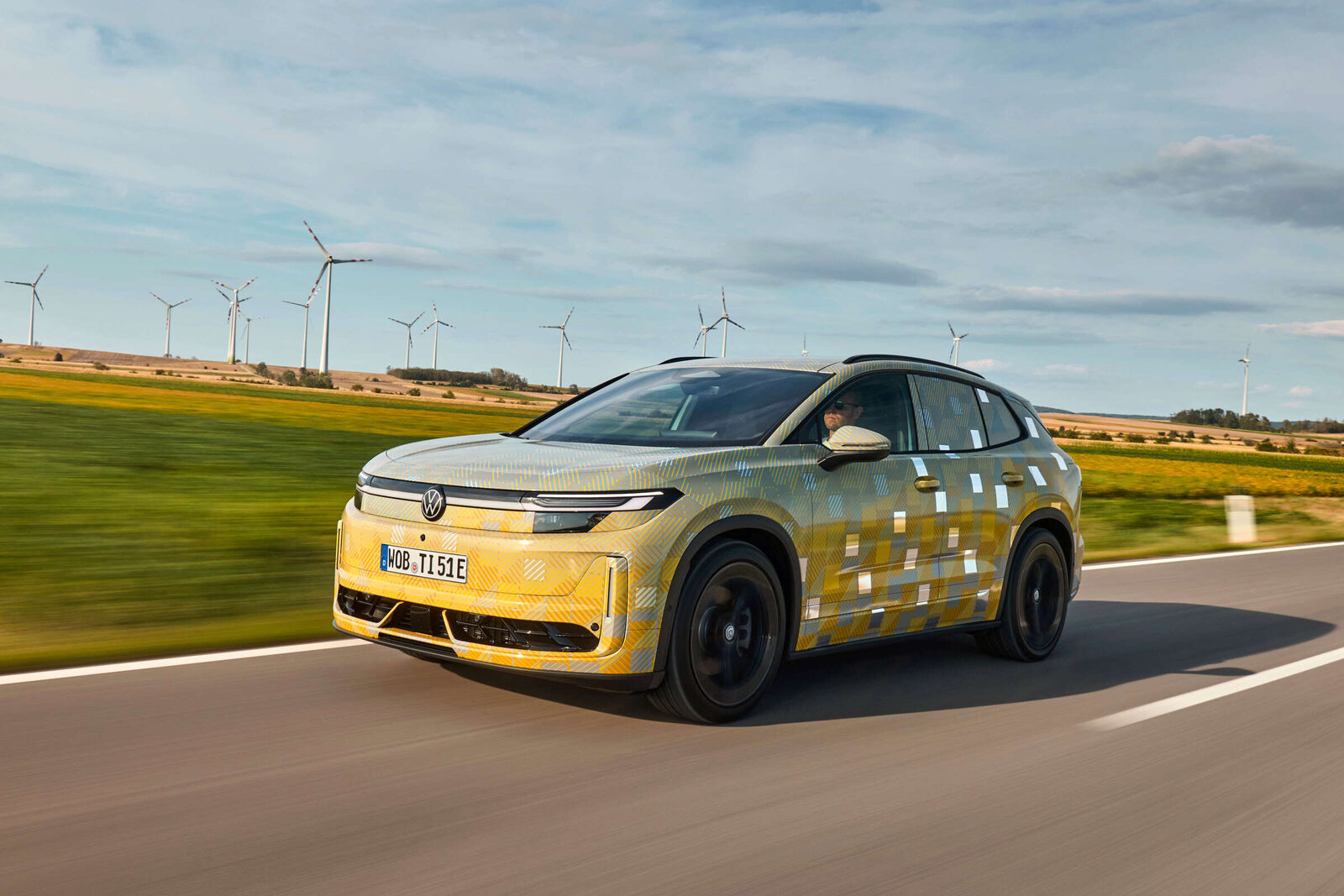 Getarnter VW ID. Tiguan mit gelb-grauer Folierung fährt auf einer Landstraße, im Hintergrund Windräder
