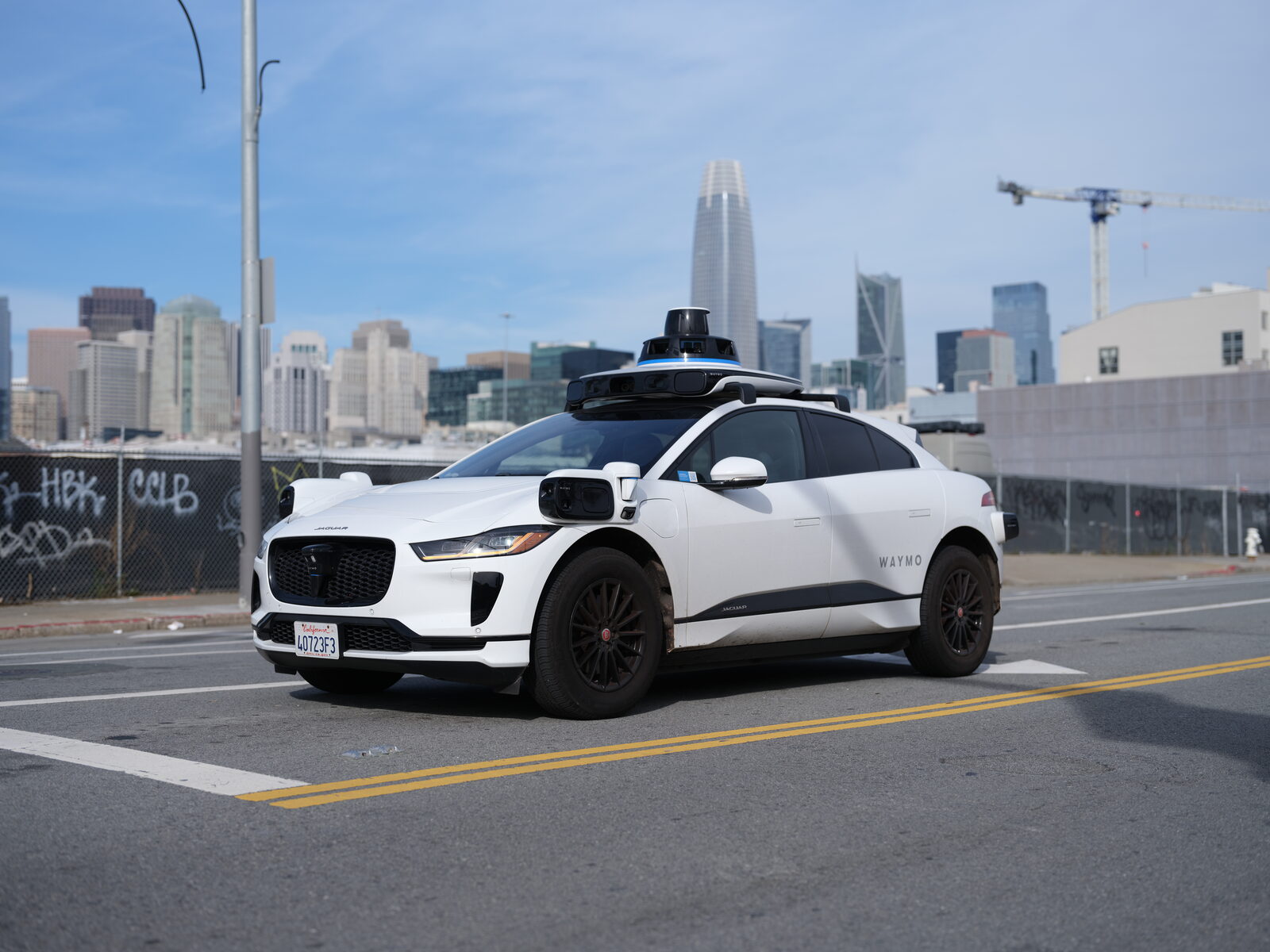 Tavanında sensör kulesi bulunan beyaz bir Waymo Jaguar I-Pace robotaksi, arka planda San Francisco gökdelenleri