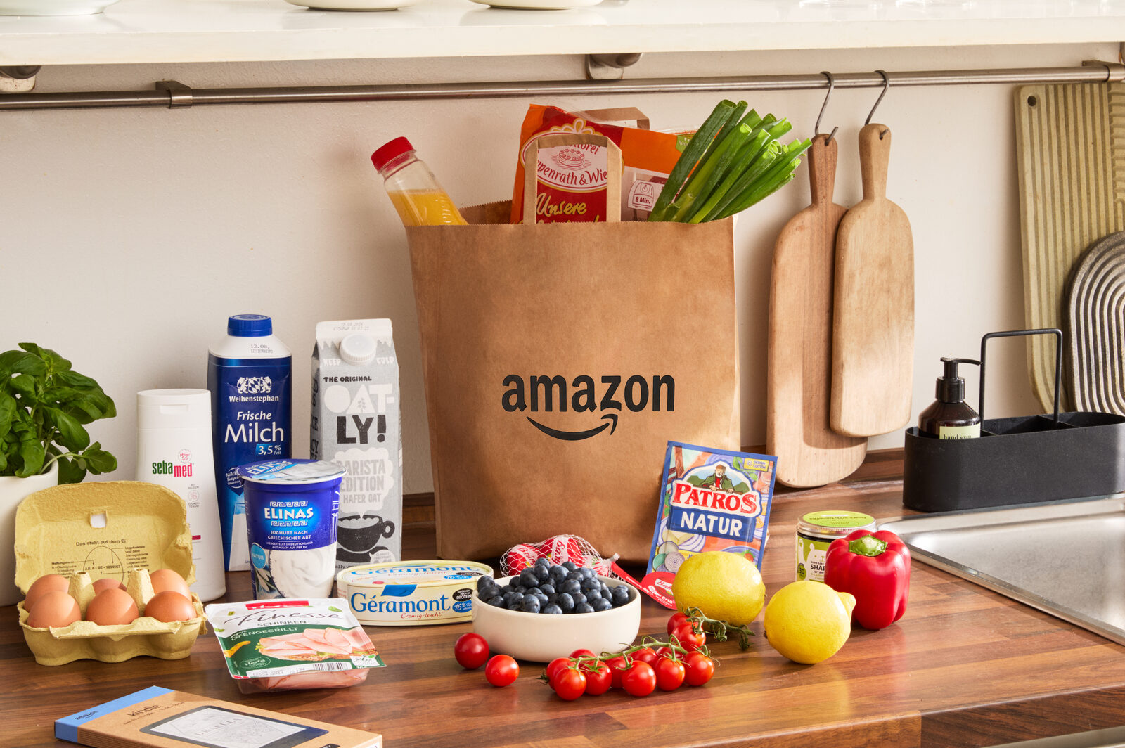 Papiertüte mit Amazon-Logo auf einer Küchenarbeitsplatte, davor Milch, Eier, Tomaten, Heidelbeeren und Käse