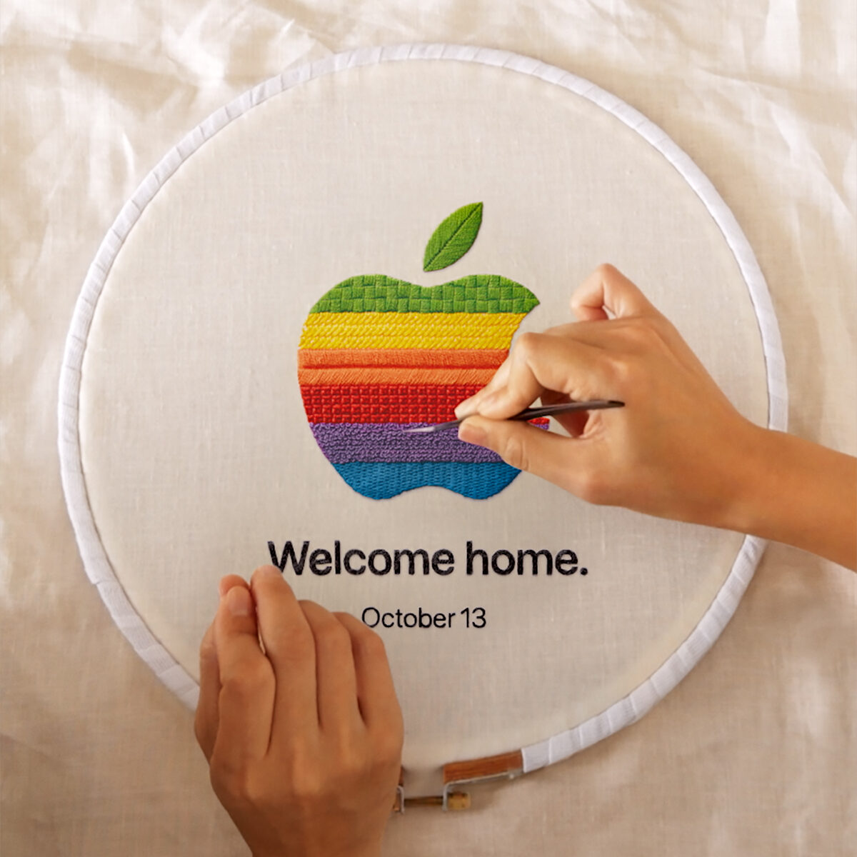 Kumaş üzerine gökkuşağı renkli Apple logosu işlenen kasnak ve altında "Welcome home. October 13" yazısı