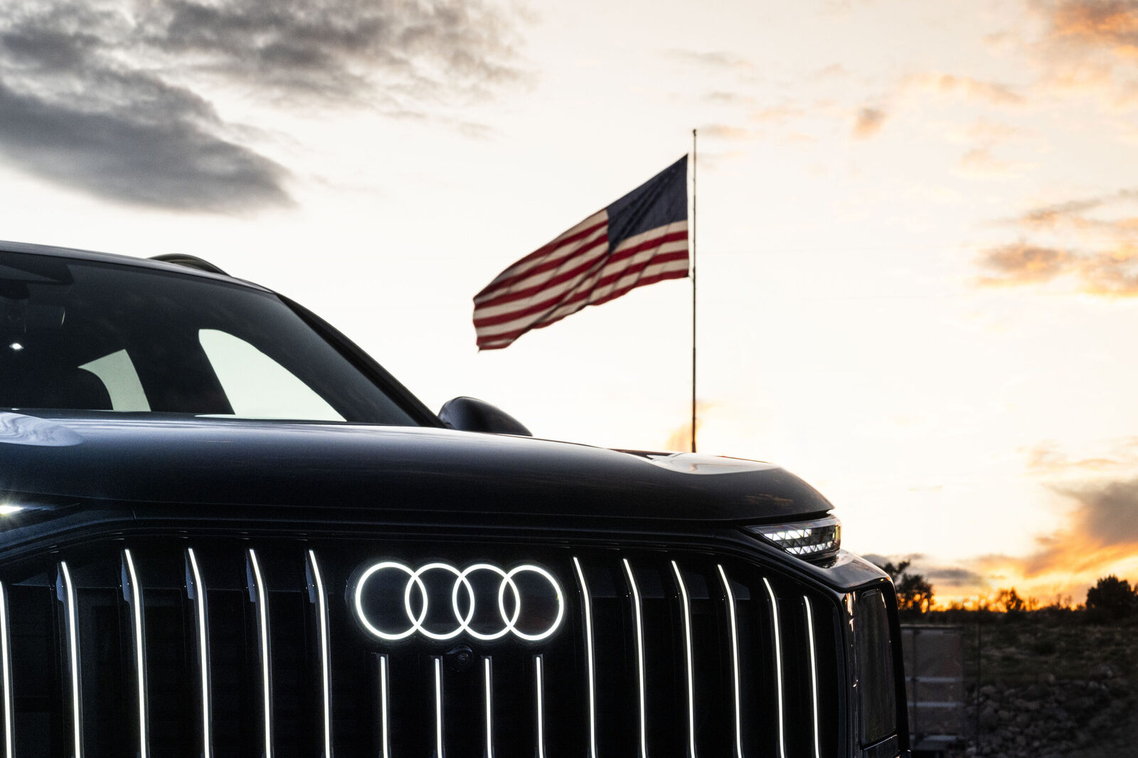 Beleuchtete Audi-Ringe am Kühlergrill eines dunklen SUV, dahinter eine US-Flagge vor Abendhimmel