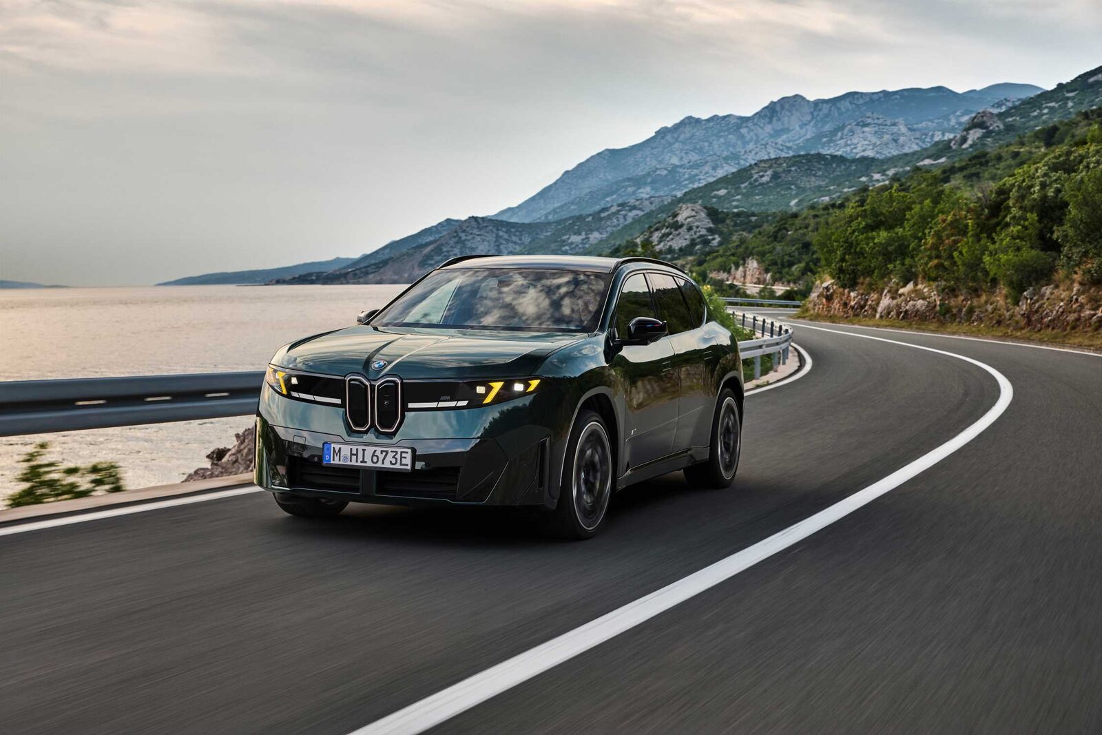 Koyu yeşil BMW iX3 M60 xDrive'ın sahil yolunda virajı dönerken önden görünümü
