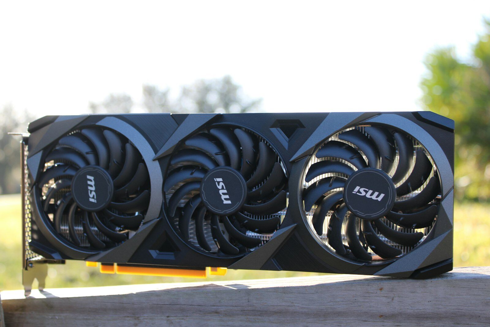 Grafikkarte mit drei Lüftern und freiliegender PCIe-Kontaktleiste auf einem Holzbalken im Freien