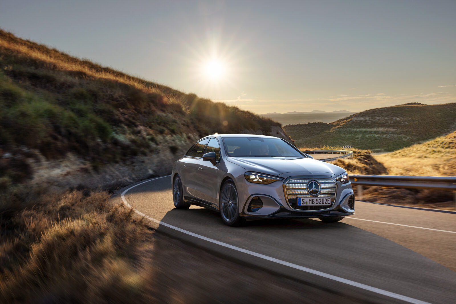 Gümüş renkli elektrikli Mercedes C-Serisi sedan, gün batımında virajlı bir dağ yolunda önden görünüm