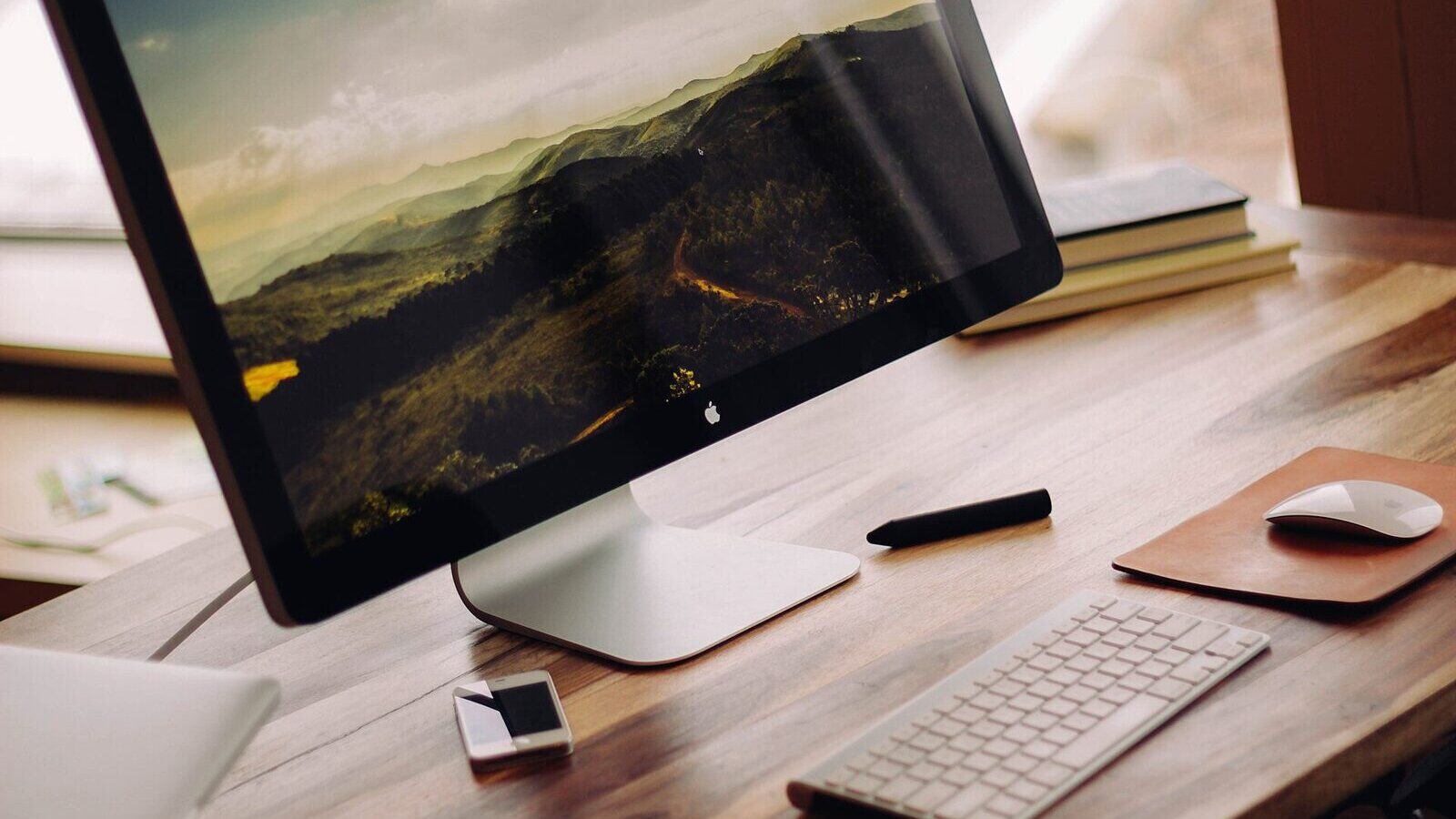 Monitor mit Landschaftsmotiv auf einem Holzschreibtisch, davor Tastatur, Maus und Smartphone