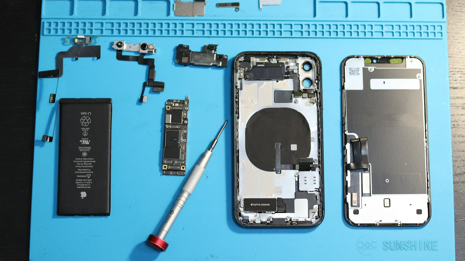 Batteria, schermo e scheda logica sono i ricambi più richiesti. Immagine: Unsplash / Fotografia Lui Vlad Componenti di un iPhone smontato su un tappetino da riparazione blu, con batteria, scheda logica e cacciavite