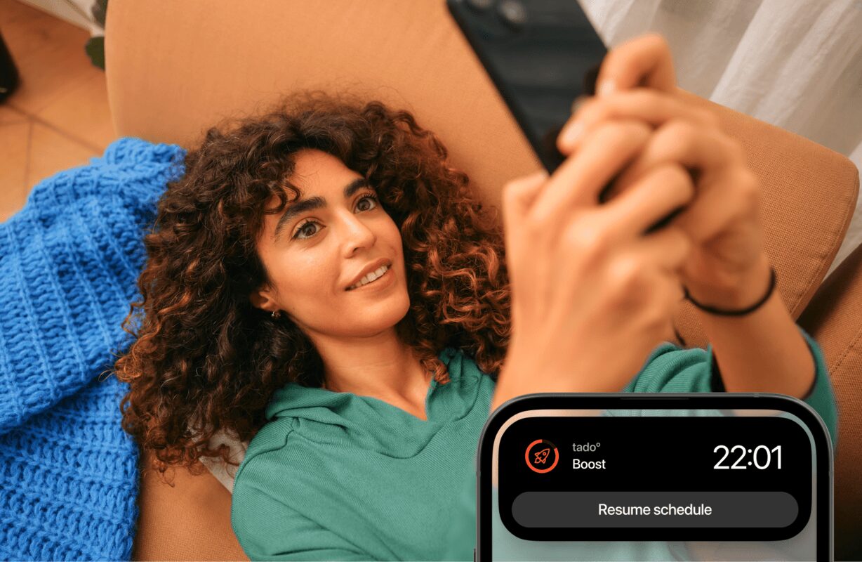Frau auf dem Sofa mit Smartphone, eingeblendet die tado° Live-Aktivität „Boost“ mit Uhrzeit und Schaltfläche zum Fortsetzen des Zeitplans