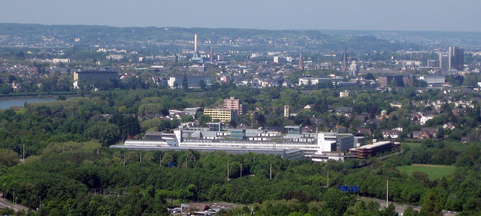 Luftbild des Telekom-Campus in Bonn-Beuel, ein langgestreckter weißer Gebäudekomplex zwischen Bäumen, dahinter die Stadt Bonn