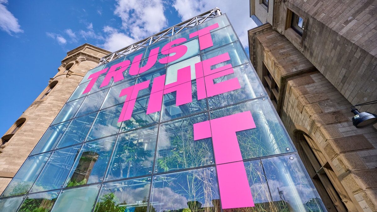 Gläserne Fassade mit dem pinkfarbenen Schriftzug „Trust the T“ zwischen zwei Sandsteingebäuden, von unten fotografiert