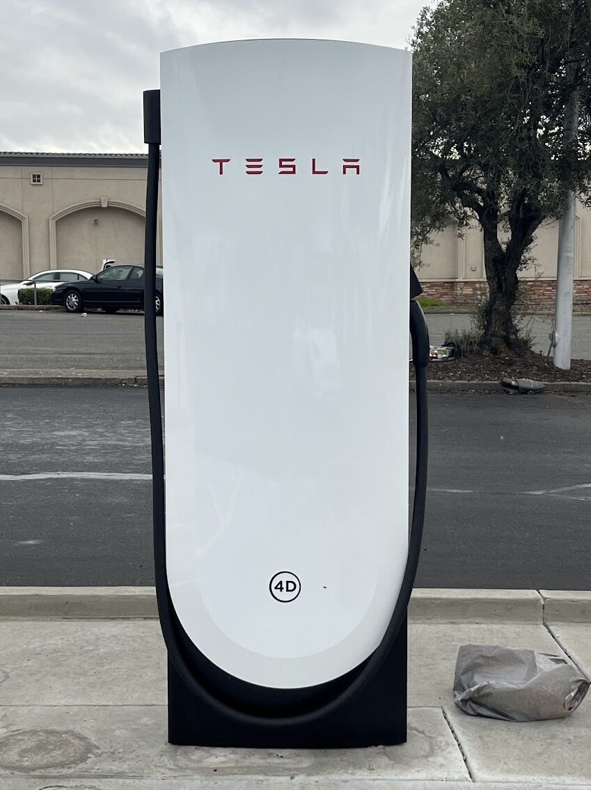 Beyaz gövdeli, üzerinde kırmızı Tesla yazısı bulunan V4 Supercharger şarj ünitesi ve siyah şarj kablosu