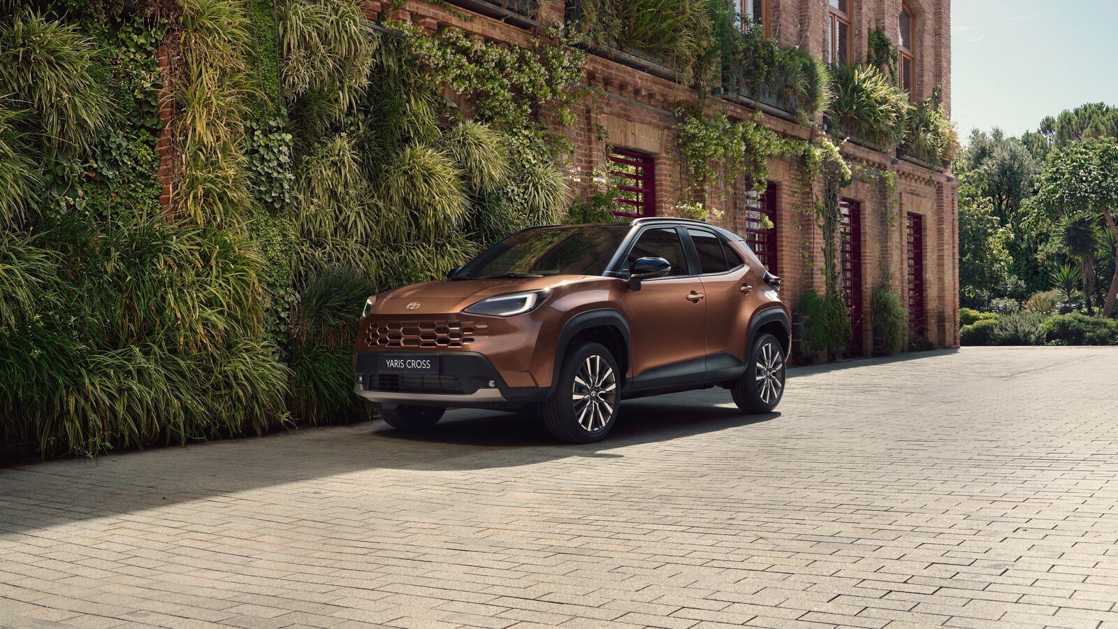 Bronz renkli yenilenen Toyota Yaris Cross Hybrid, sarmaşıklı tuğla bir binanın önünde önden çapraz açıyla duruyor