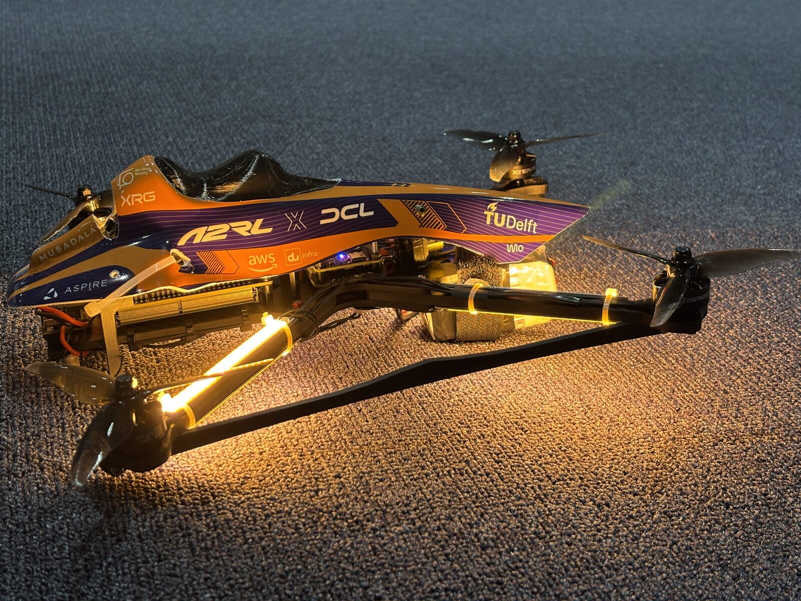 De racedrone van TU Delft met paars-oranje behuizing en verlichte armen op een tapijtvloer
