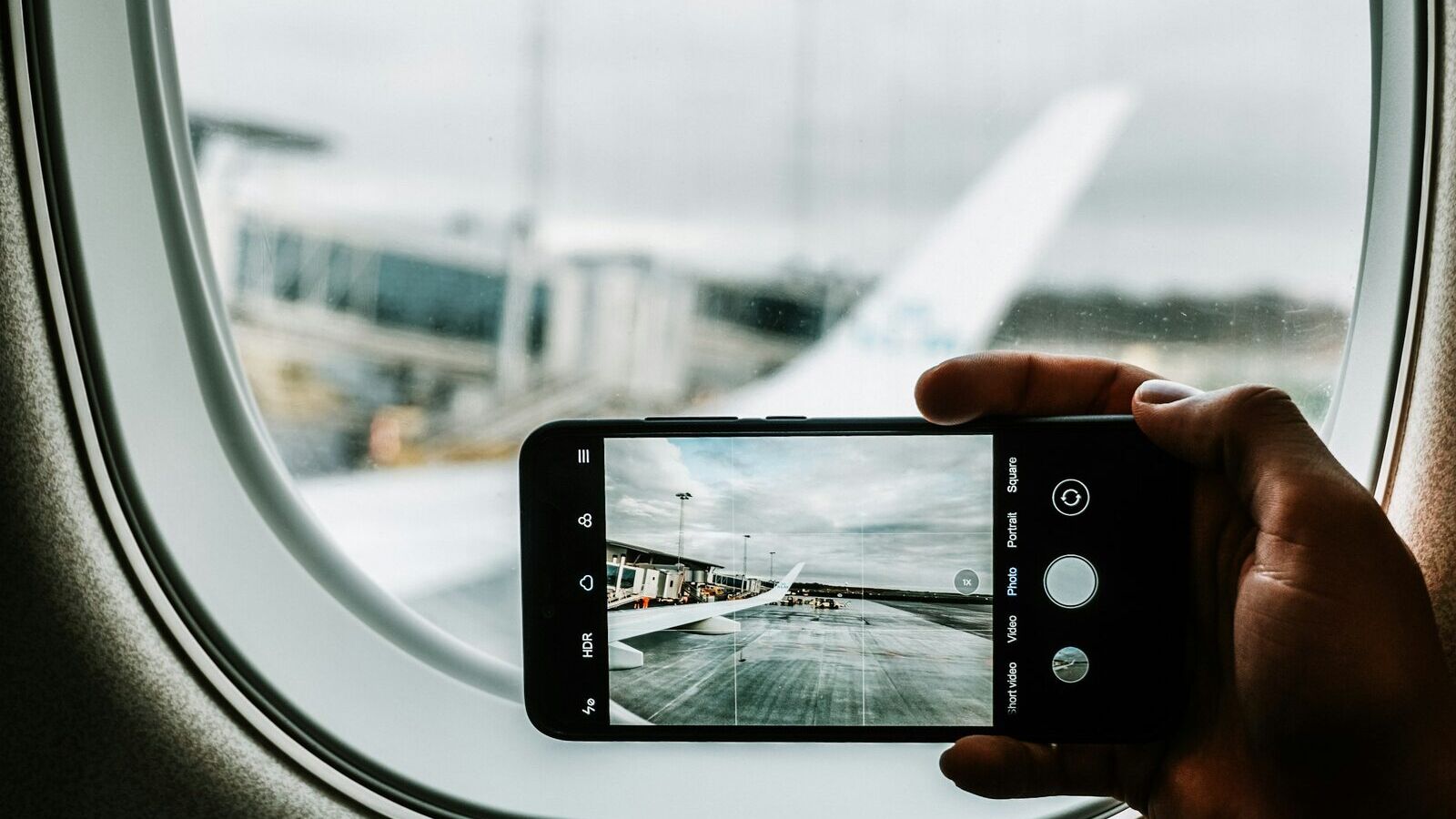 Uçak penceresinin önünde havalimanı apronunu fotoğraflayan akıllı telefon